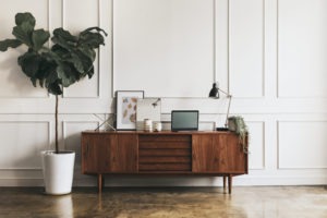 Mid-century modern wooden dresser against a white wall with wood detailing. there are items on the dresser including a lamp and pictures. There is also a plant in front of the dresser